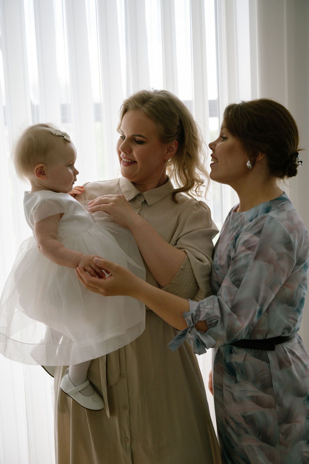 Two women in elegant dresses admire a baby girl in a white dress near a bright window.