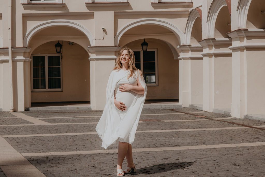 Pregnant woman in white dress posing outdoors near arched building on sunny day.