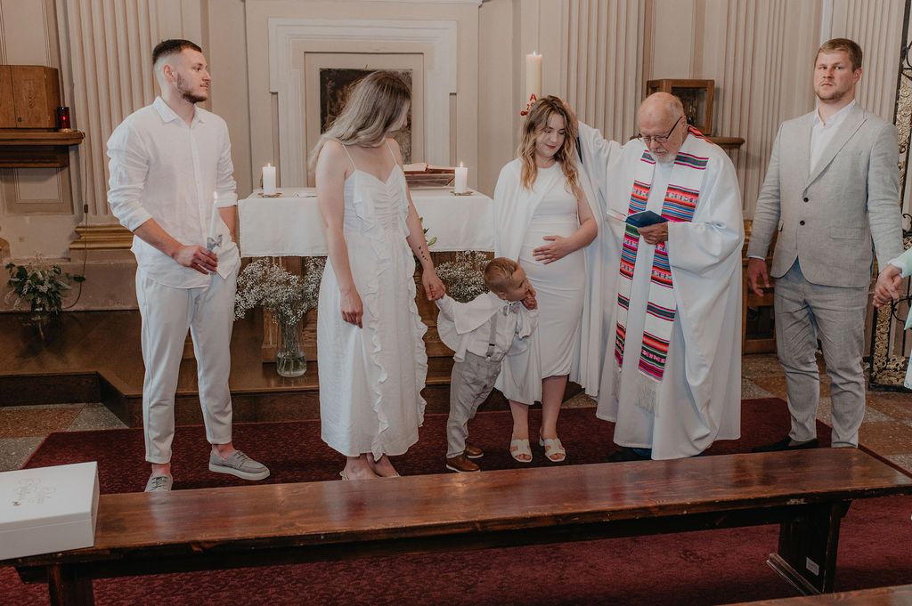 Wedding ceremony in a church with couple in white, officiant in colorful stole, and guests standing at altar.