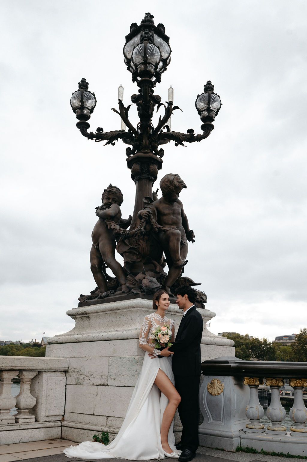 Bride and groom pose by ornate Pont Alexandre III lamp post in Paris, bride holding bouquet in elegant white gown.