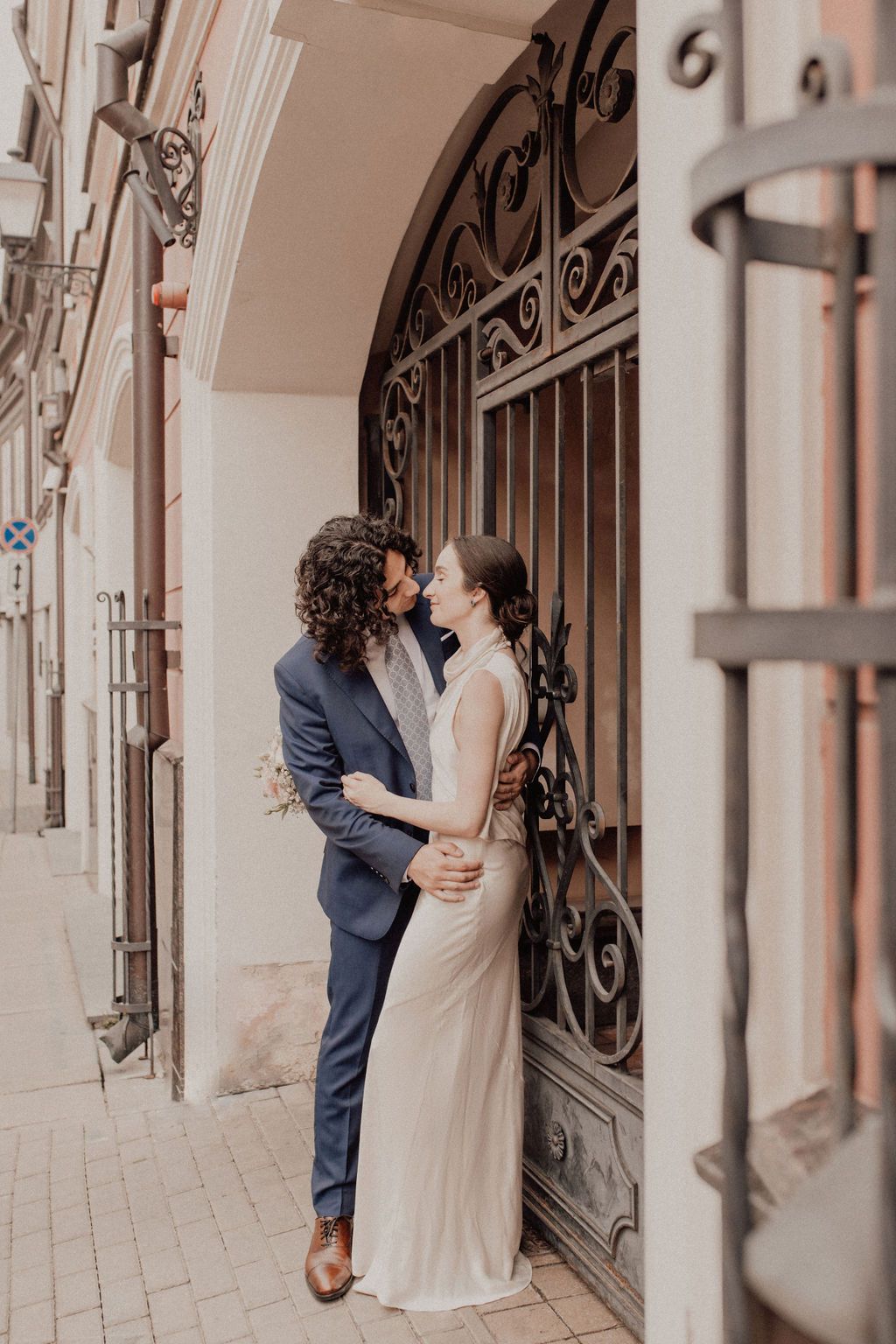 Couple in wedding attire embracing by ornate iron gate on cobblestone street, groom in navy suit, bride in white gown.