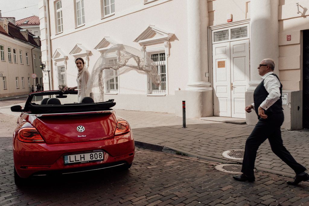 Bride in wedding dress standing on red VW Beetle convertible on cobblestone street in European city.