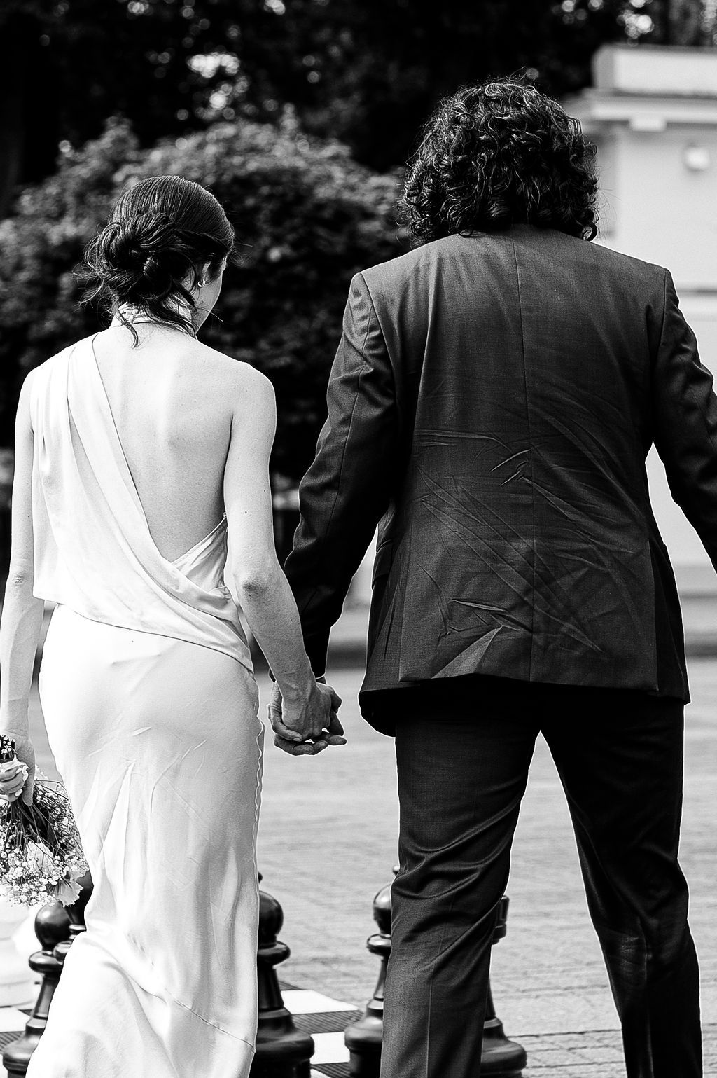 Bride and groom walk hand in hand, black and white photo showing elegant backless wedding dress.