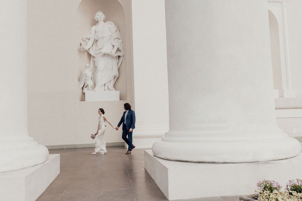 Bride and groom walking hand in hand past white marble columns and a classical statue in a grand hall.