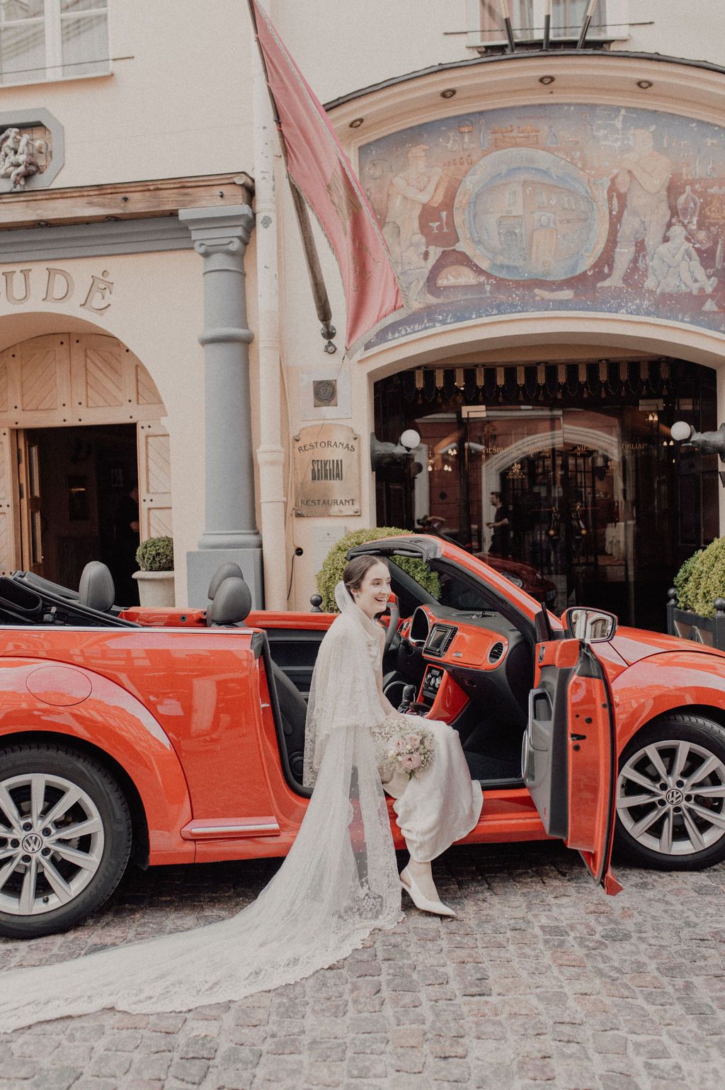 Elegant bride in white gown with long veil posing beside red convertible outside a European hotel.