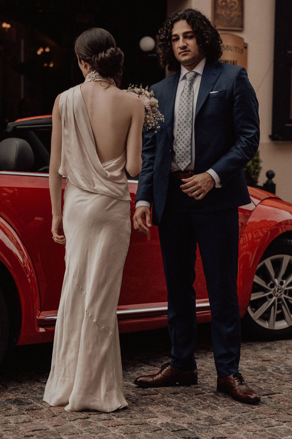 Elegant couple in wedding attire posing beside a red car, bride in backless gown, groom in navy suit.