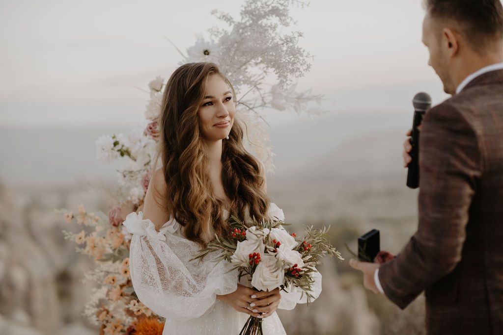 Beautiful bride with long wavy hair holding a colorful bouquet smiling during an outdoor mountaintop ceremony.