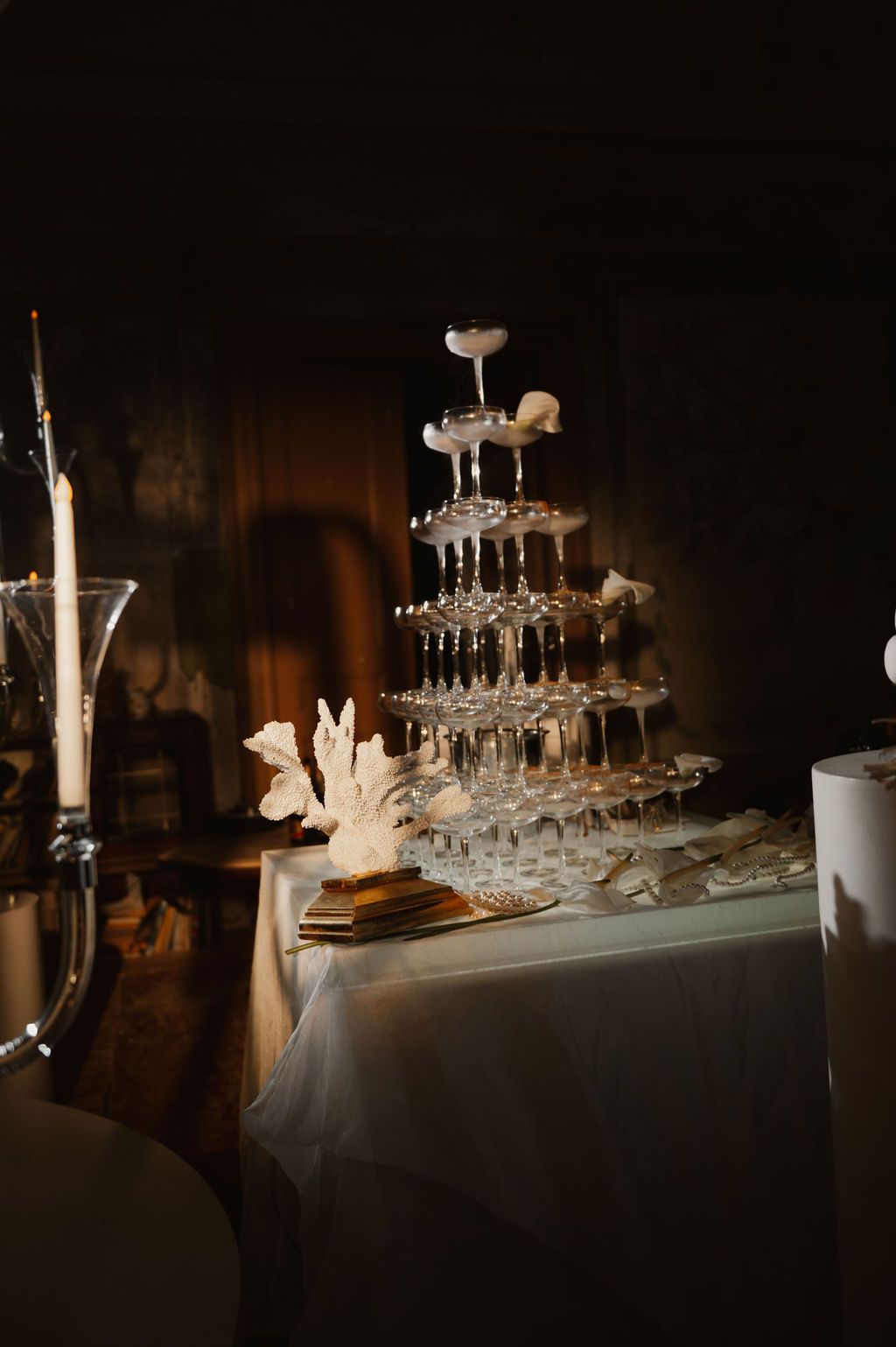 Elegant champagne tower with crystal glasses on a white draped table, adorned with coral decor and candlelight.