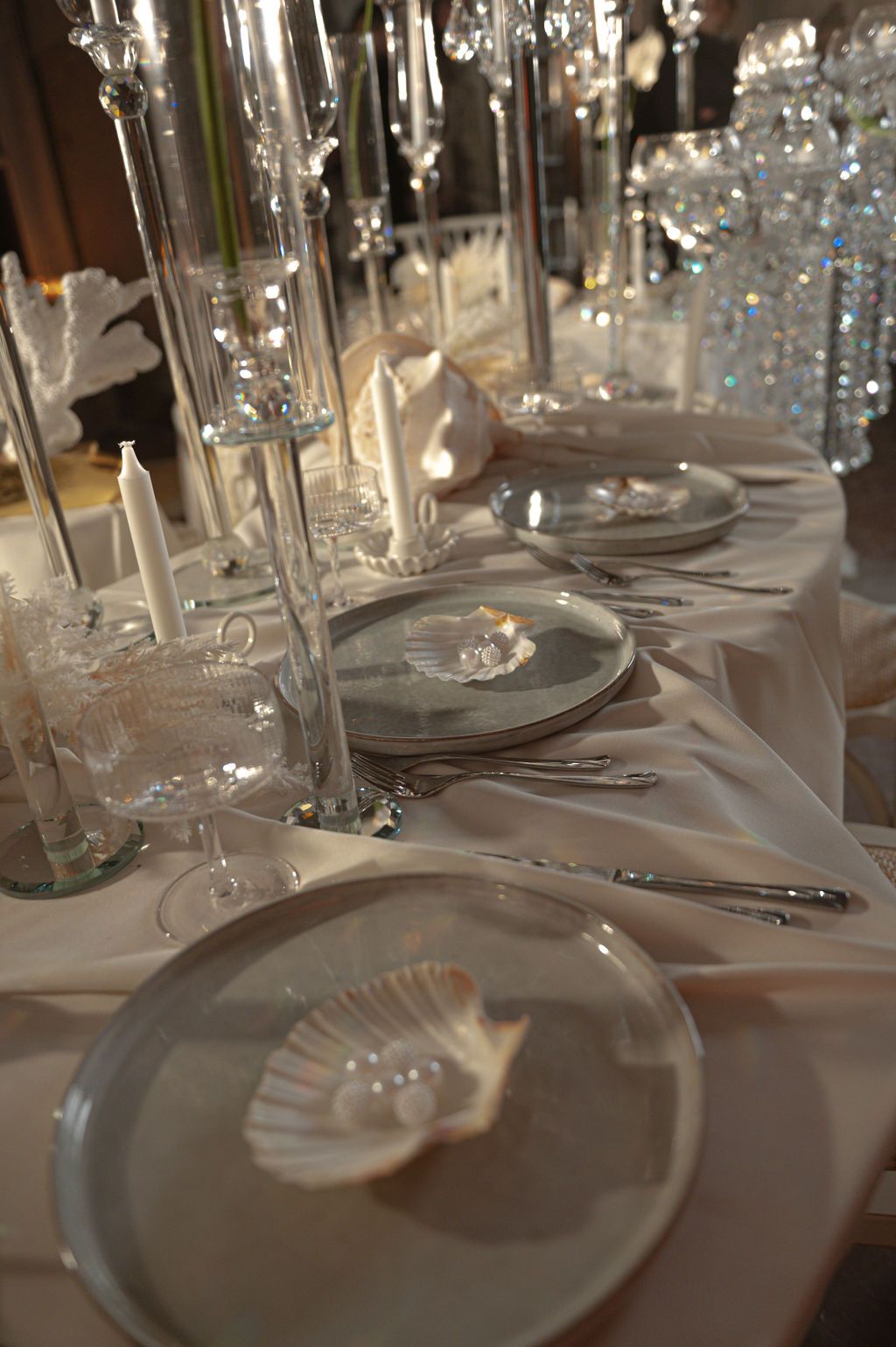 Elegant wedding table setting with crystal candleholders, white plates, seashell accents, and silver cutlery on a white tablecloth.