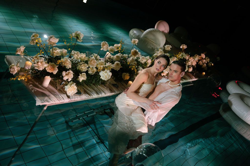 Couple in wedding attire relaxing by a pool decorated with elegant floral arrangements and soft lighting.