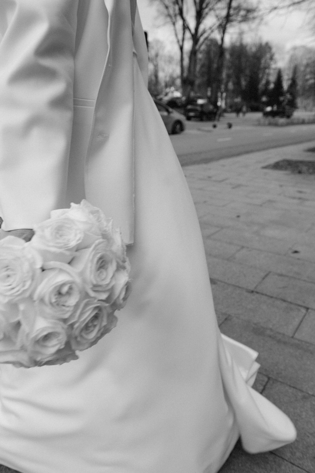 Black and white photo of a bride holding a round rose bouquet beside her flowing wedding gown on a cobblestone path.