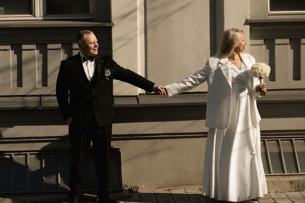 Bride in white suit and groom in black tuxedo hold hands outdoors on their wedding day.
