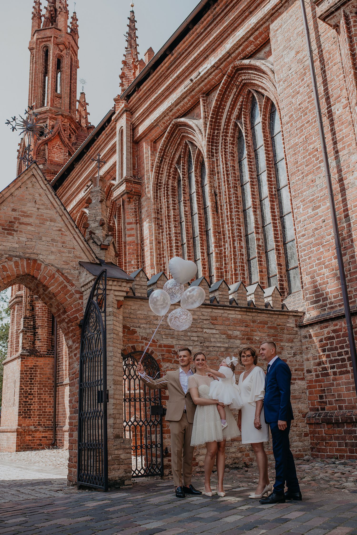 A group in formal attire poses in front of a Gothic-style red brick church with ornate spires and arches.