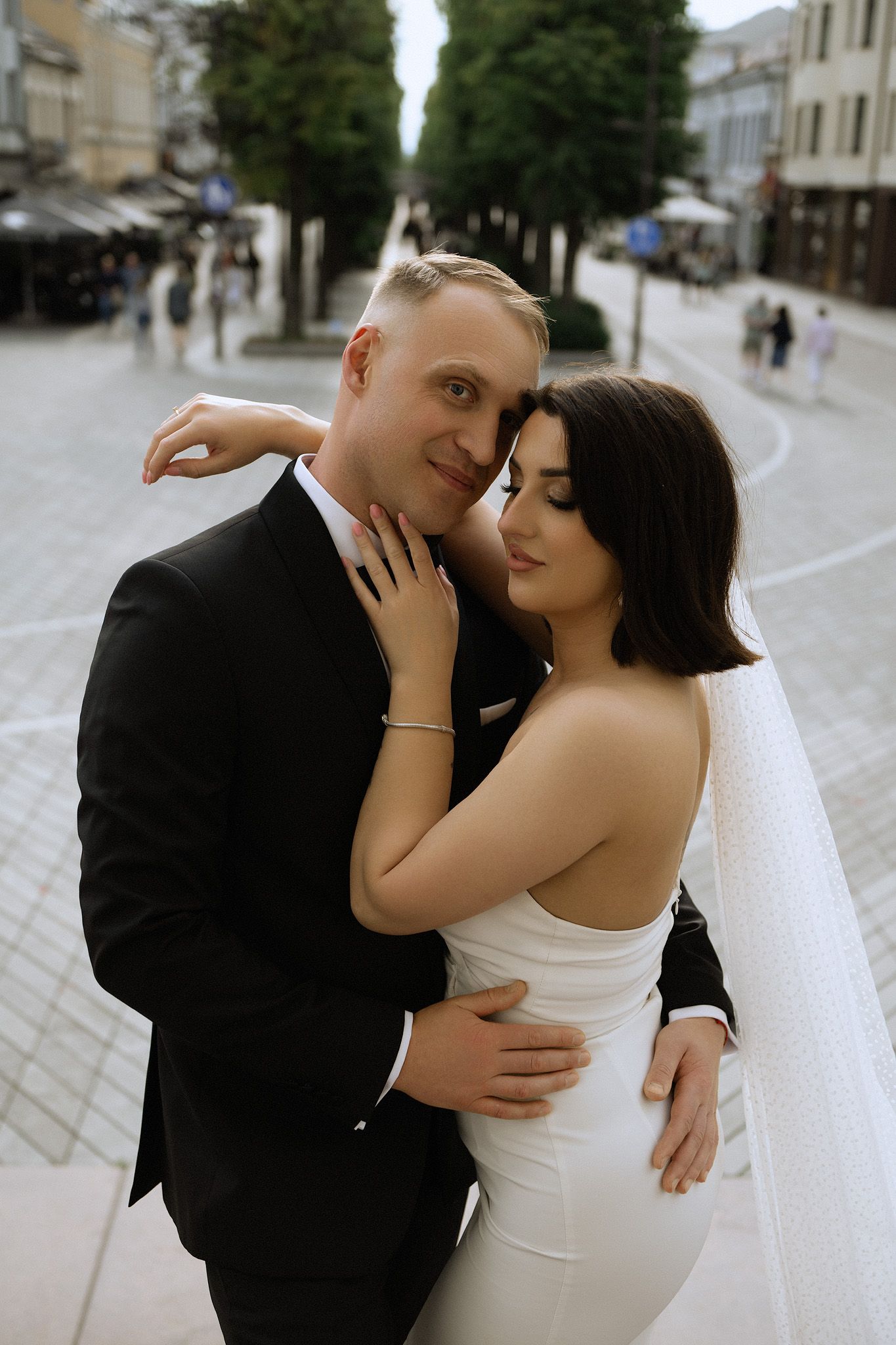 A romantic wedding couple embraces on a quiet European cobblestone street lined with trees and buildings.