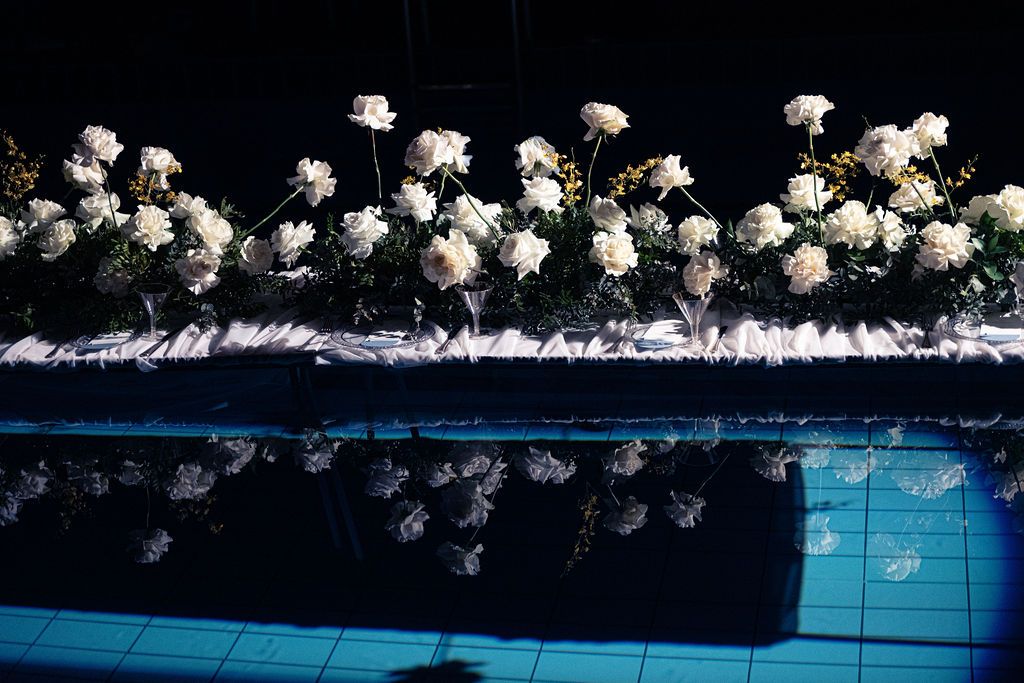White roses arranged along a reflective poolside display, mirrored in the blue water below.