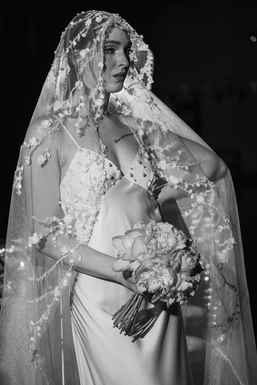 Elegant bride in a satin gown wearing an ornate floral lace veil holding a peony bouquet.