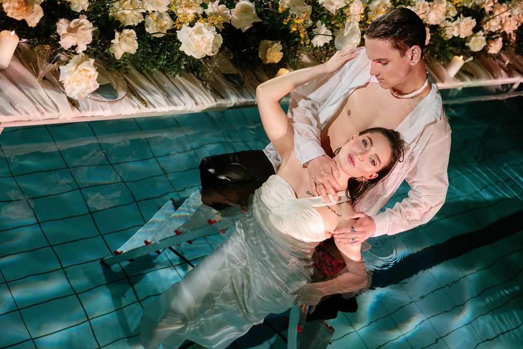 Couple in wedding attire posing romantically in a pool surrounded by floral decorations.