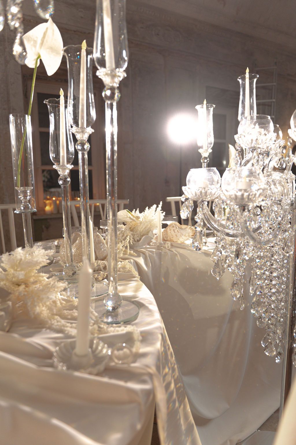 Elegant white wedding reception table with crystal candelabras, white floral centerpieces, and satin tablecloth in a grand venue.