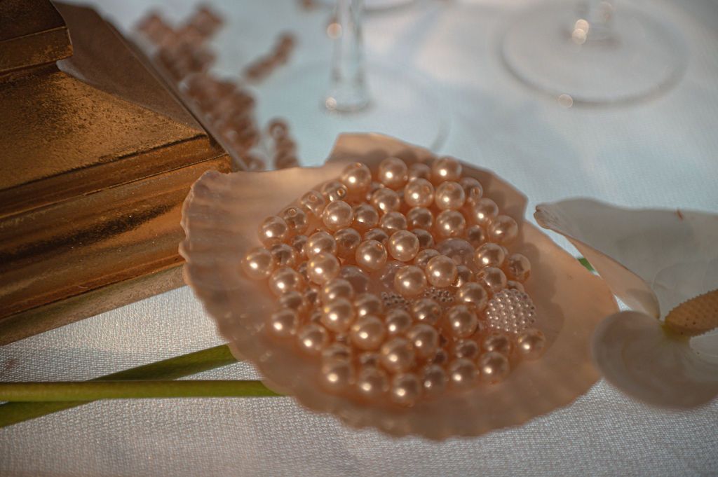 Scallop shell filled with pink pearls on white linen table with gold book and wine glass in background.