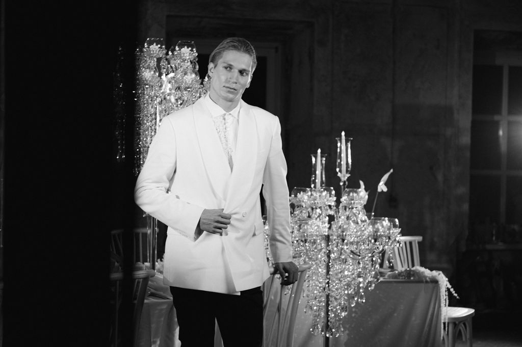 Elegant man in white suit jacket standing near crystal candelabras in a black and white photo.