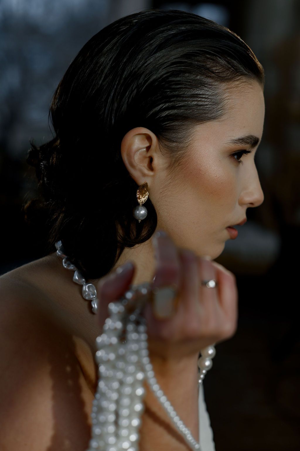 A woman in profile wearing gold pearl drop earrings and a layered pearl necklace with sleek dark hair pulled back.