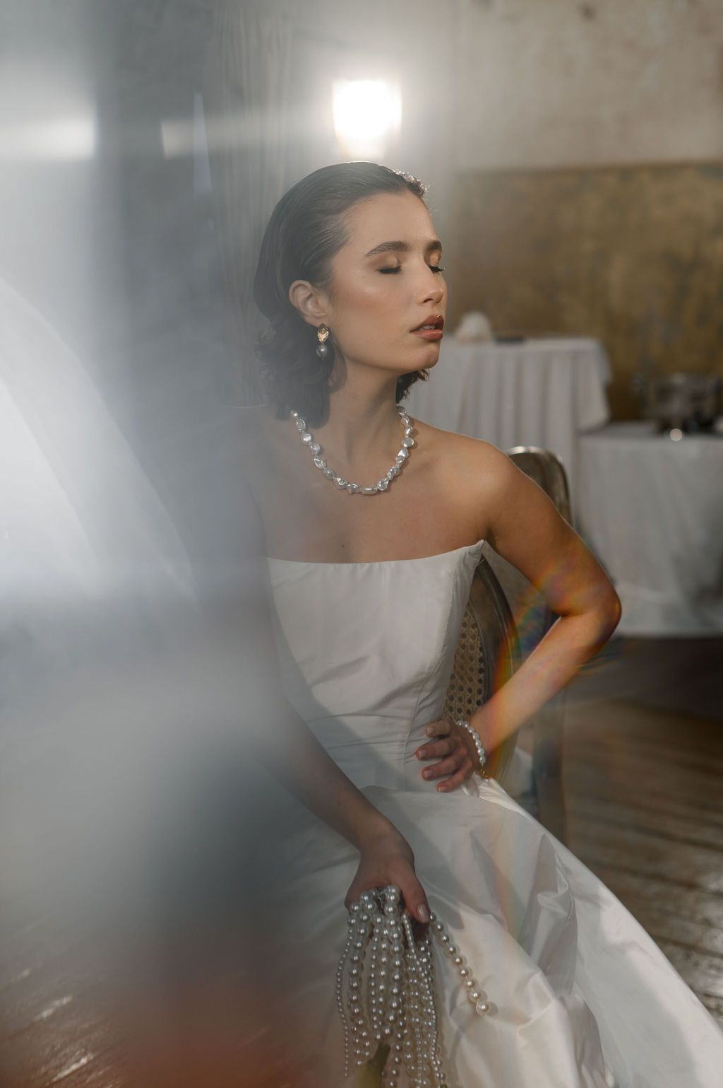 Elegant bride in strapless white gown with pearl necklace and earrings, posing gracefully in soft light.