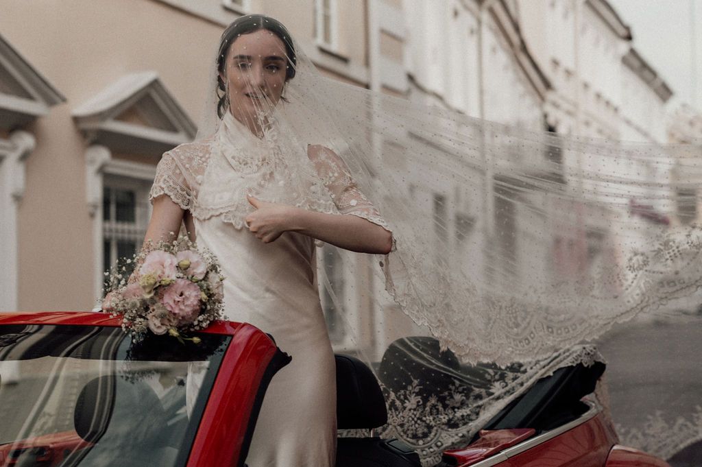 Bride in lace veil and white dress holding bouquet, leaning out of red convertible car on a city street.