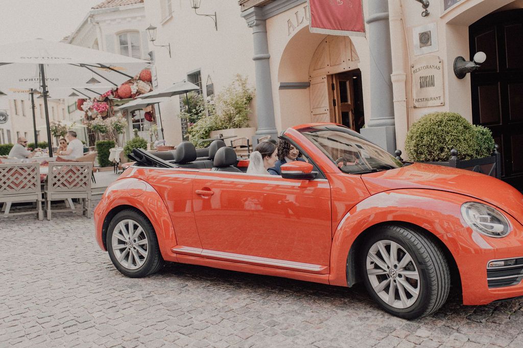 Red VW Beetle convertible parked on cobblestone street near outdoor cafe and building entrance.