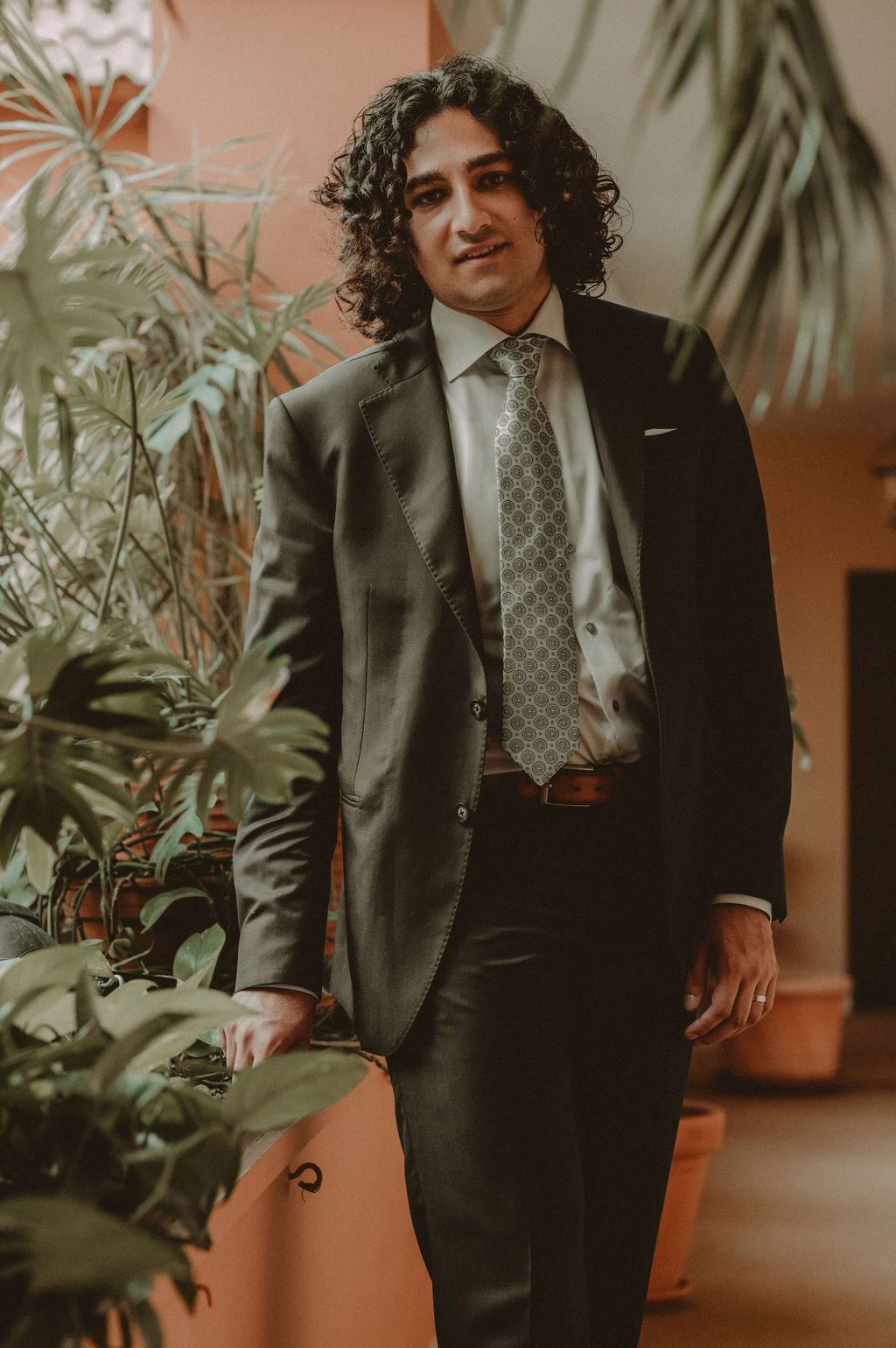 Man in dark suit and patterned tie with curly hair standing among green plants in a warm-toned interior.
