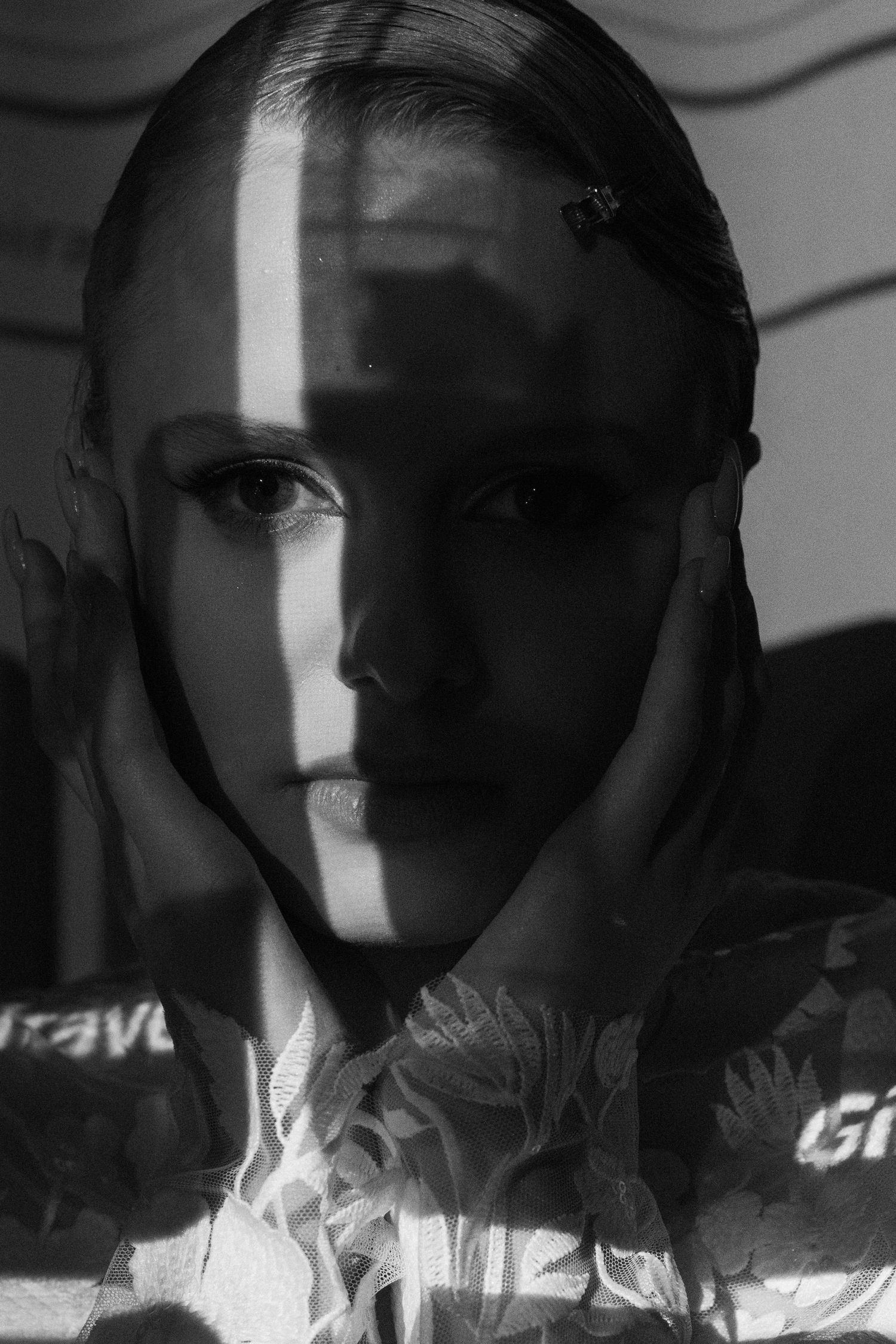 Black and white portrait of a person with dramatic shadow lighting across their face, hands framing their cheeks.