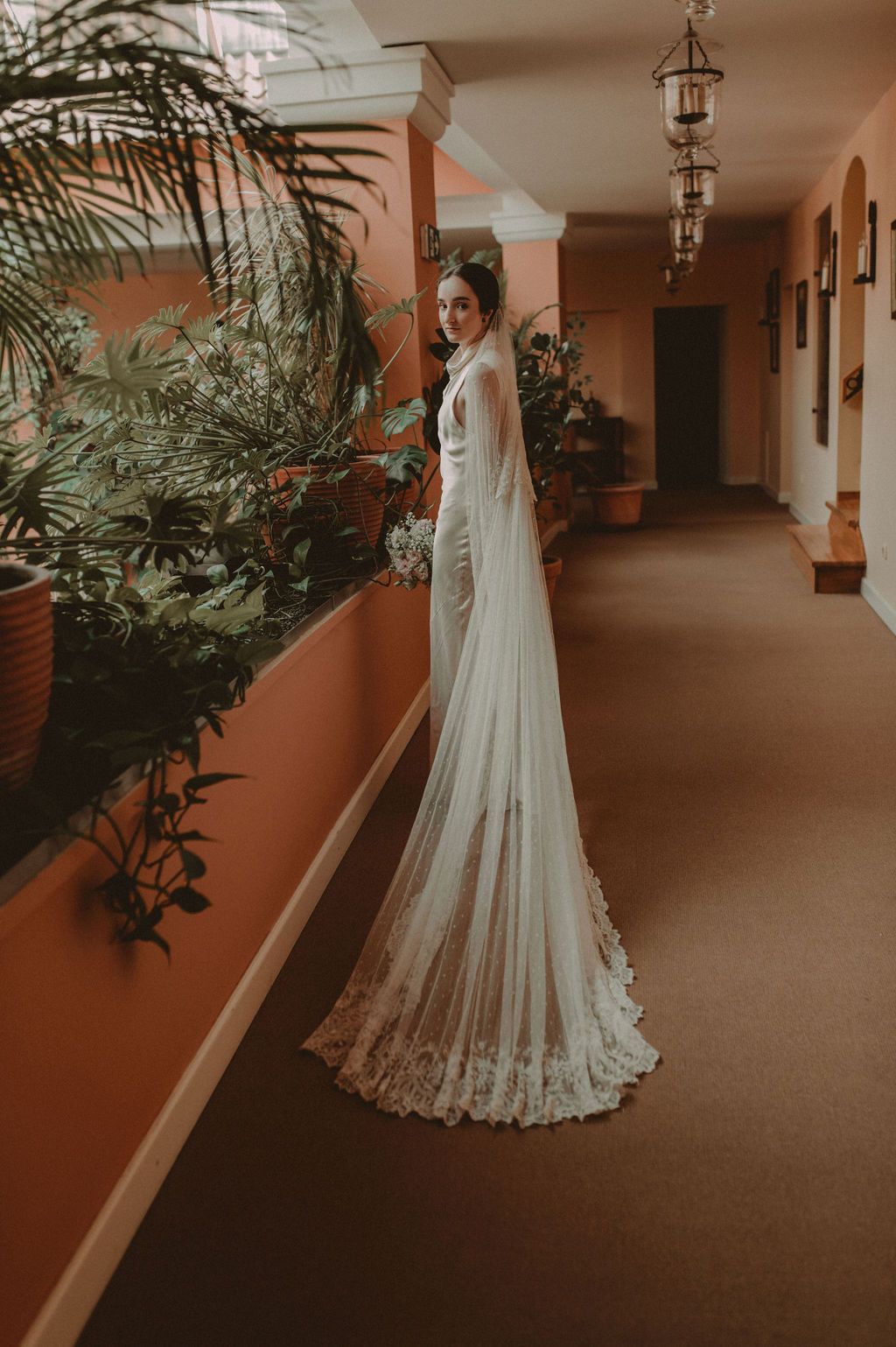 Bride in a sleek white gown with a long lace veil walking through a warm-toned hotel corridor.