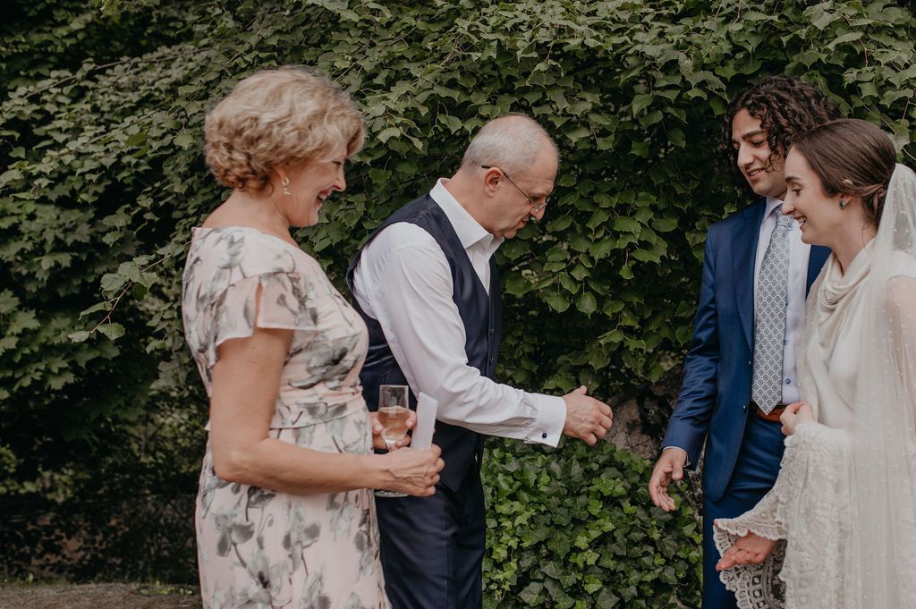 Wedding guests and groom in vest socializing outdoors near lush green ivy wall.