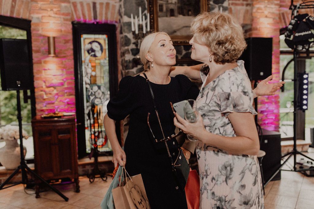 Two women greeting each other warmly at an indoor event, holding shopping bags in a decorated venue.