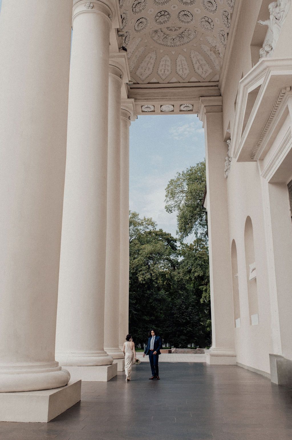 Couple holding hands between grand white columns of a neoclassical building with ornate ceiling and lush green trees.
