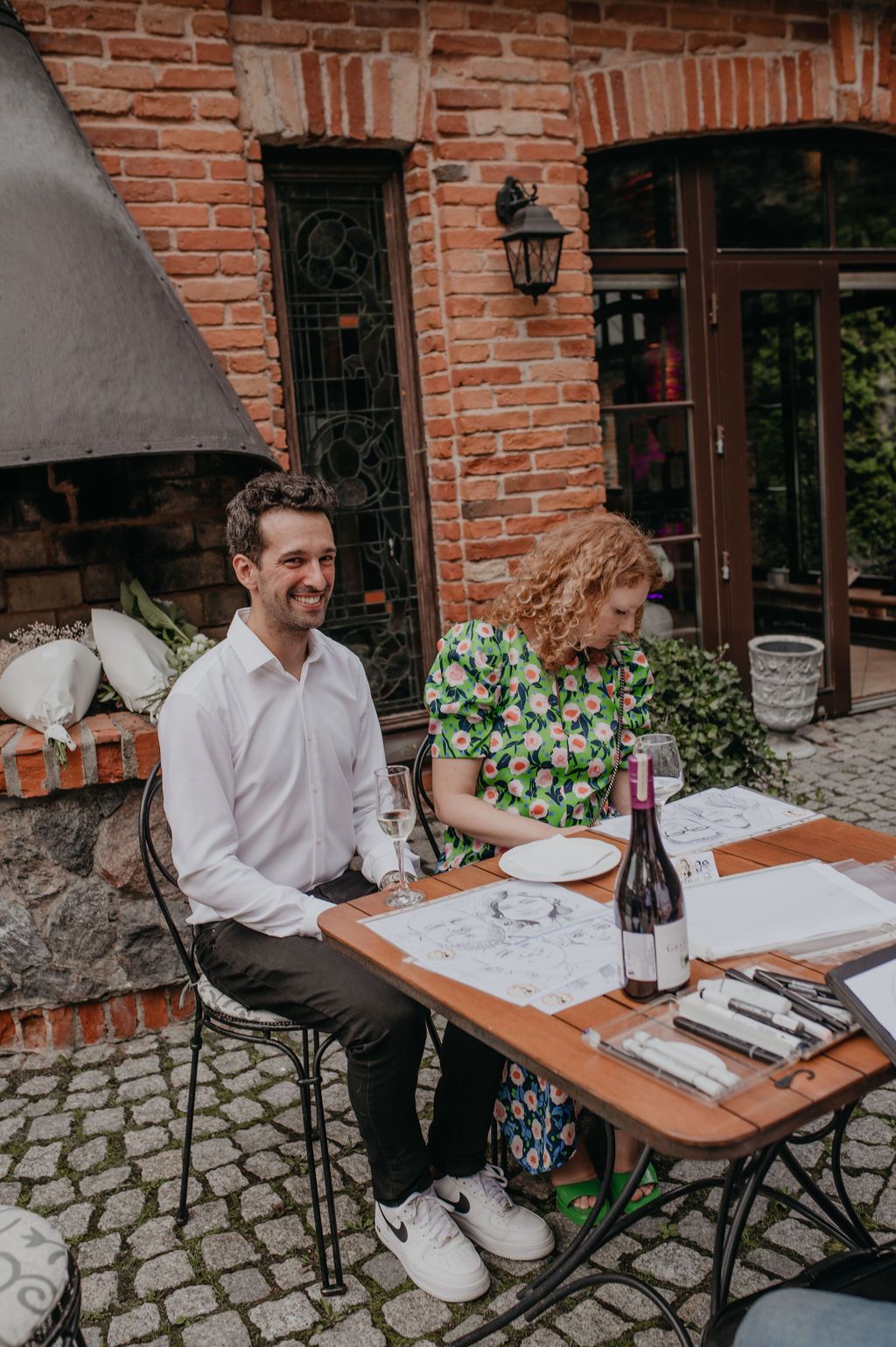 Couple dining outdoors at a rustic brick restaurant patio, with wine bottle on table, man in white shirt smiling.