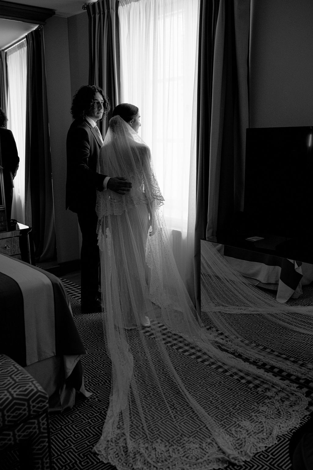 Black and white silhouette of a bride and groom standing by a window, her lace veil trailing on the floor.
