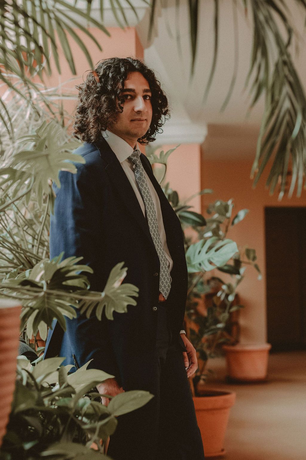 Young man with curly hair in a navy suit and tie standing among lush tropical potted plants indoors.