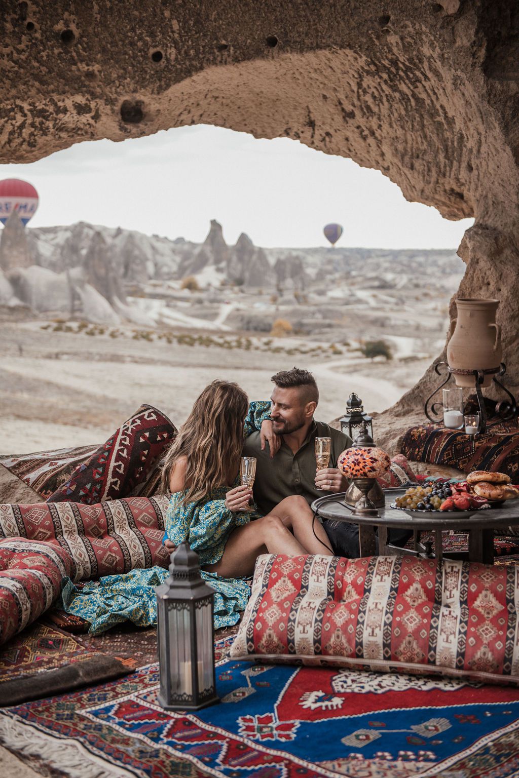 Couple enjoying a romantic cave picnic in Cappadocia with colorful rugs, lanterns, and hot air balloons in the background.
