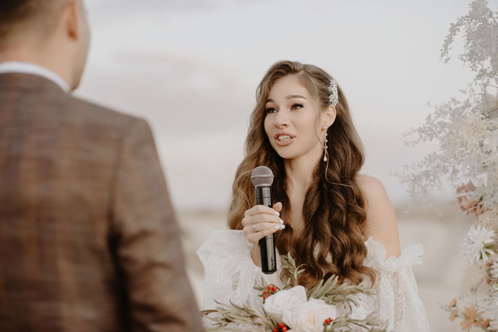 Bride with long wavy hair speaks heartfelt vows into a microphone at an outdoor wedding ceremony.