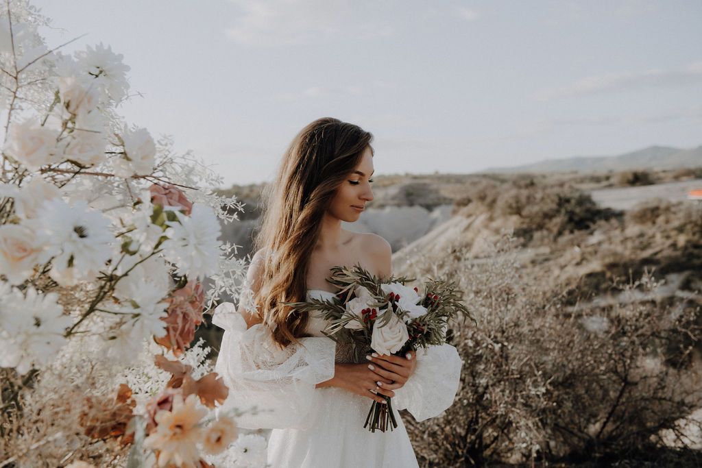 Bride in off-shoulder gown holds a white bouquet among blooming flowers in a scenic outdoor landscape.