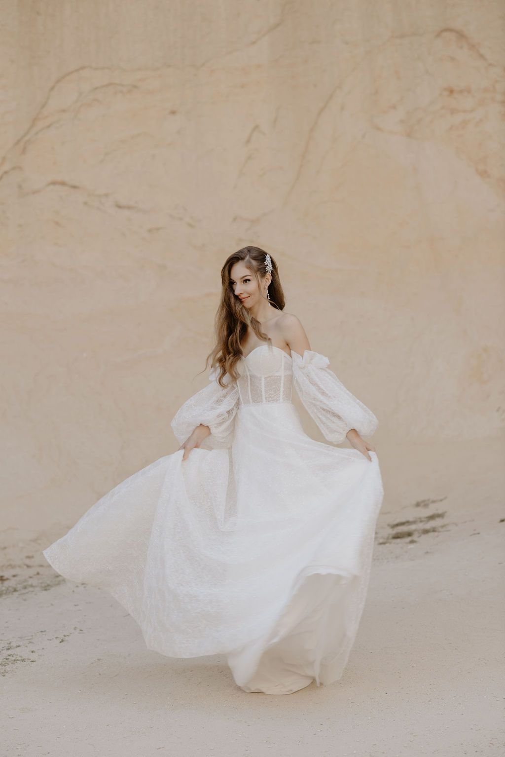 Woman in flowing white off-shoulder bridal gown twirling outdoors against a sandy beige stone wall.