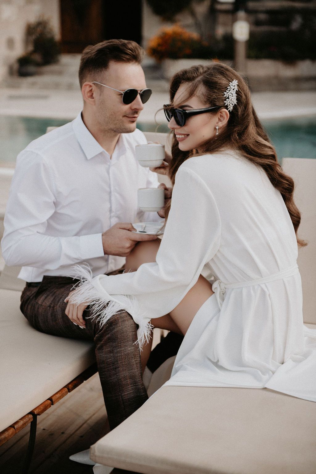 Elegant couple in white outfits and sunglasses sharing coffee by a pool, smiling romantically on a sunny day.