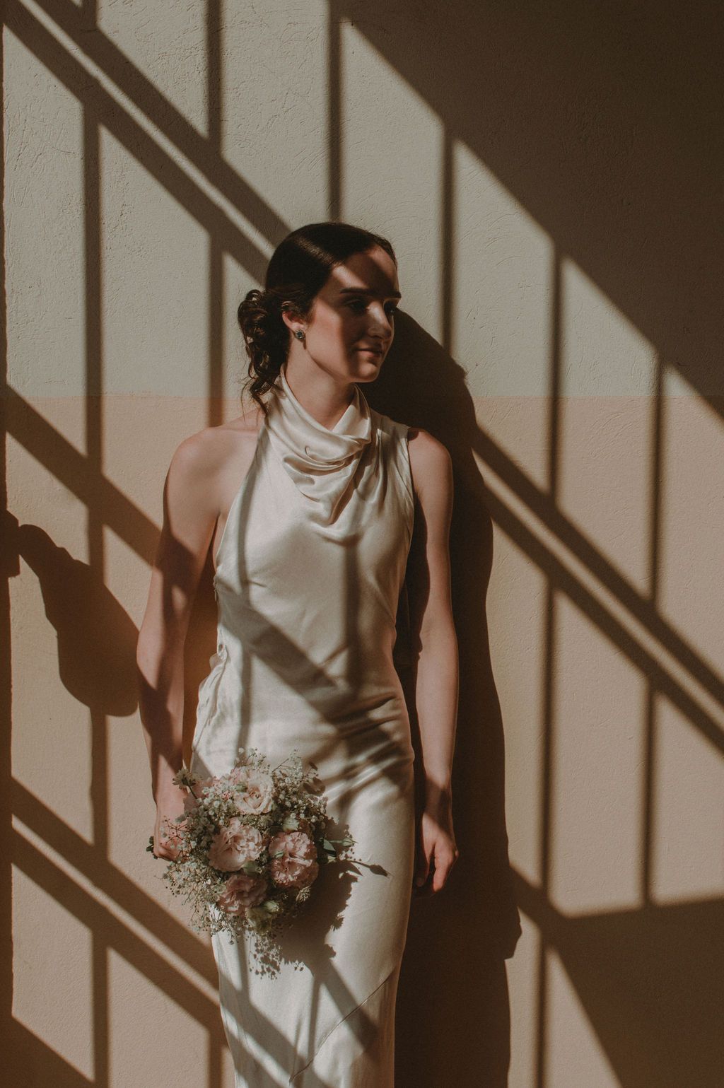 Bride in white halter gown holding pink bouquet, bathed in geometric window shadows against warm beige wall.