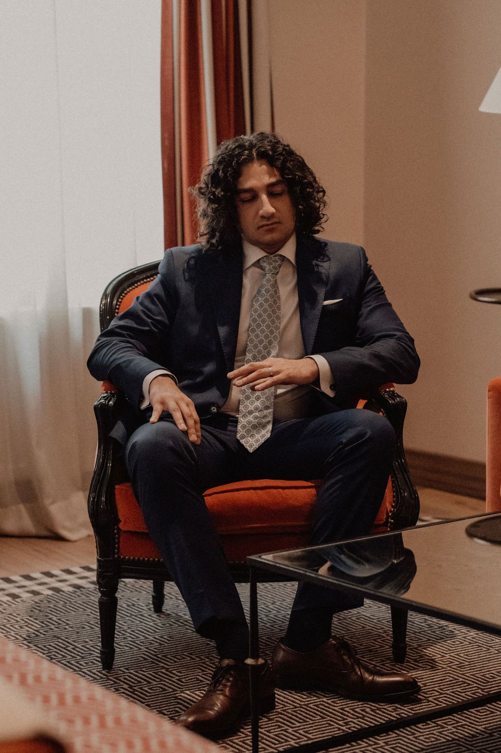A young man with curly hair sits in an ornate armchair wearing a navy suit and patterned tie.