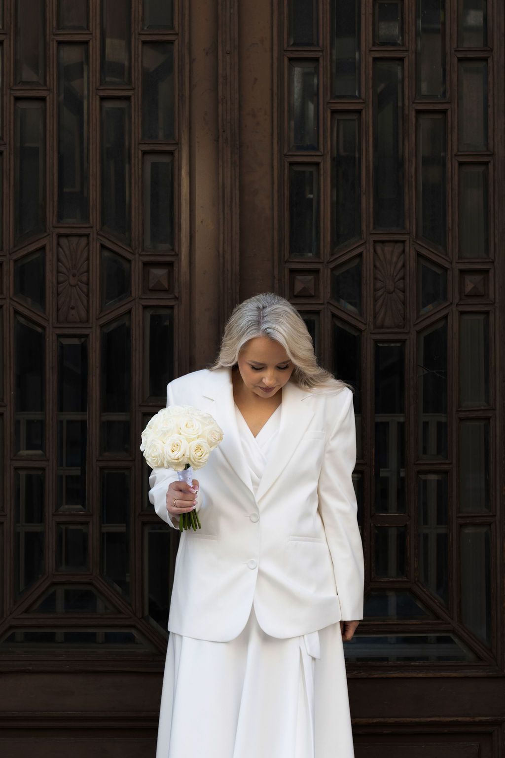 Sophisticated bride in white blazer suit looks down at white rose bouquet standing before ornate dark wooden doors.