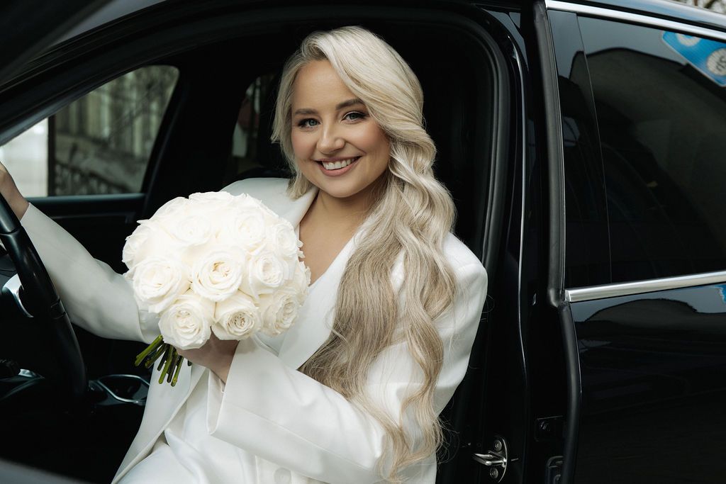 Smiling blonde bride in white dress holding bouquet of white roses while sitting in a black car.