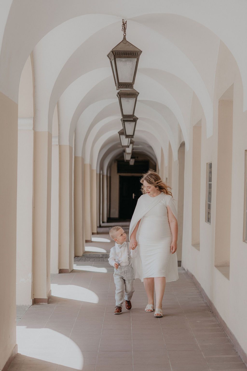 A mother in a white dress walks hand-in-hand with her toddler son through an elegant arched corridor with hanging lanterns.
