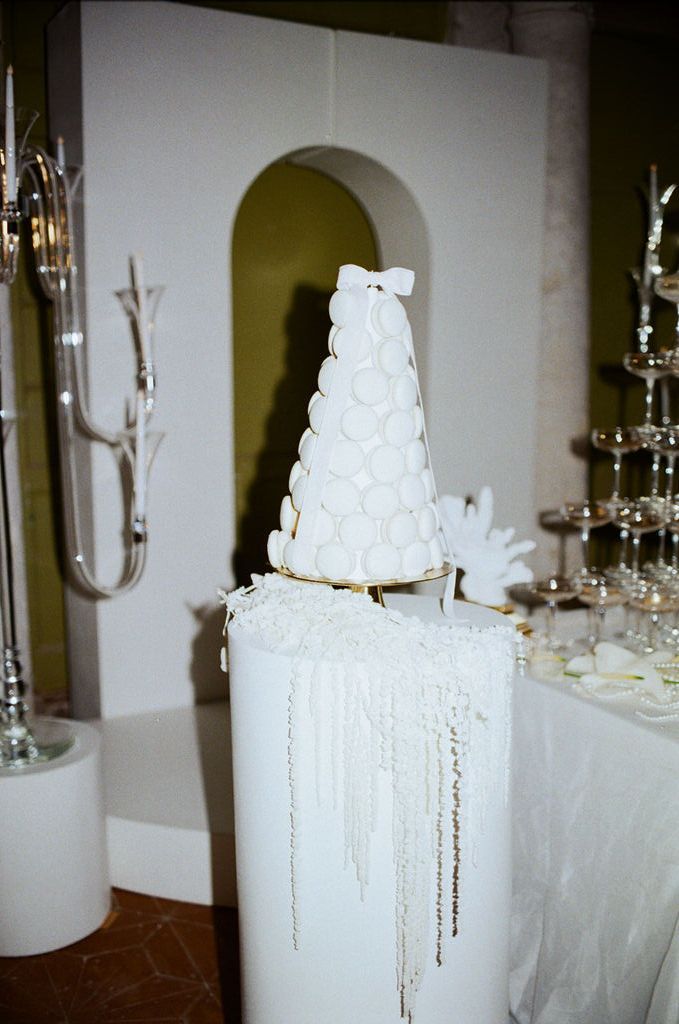 Elegant white macaron tower wedding cake on a pedestal with pearl decorations and champagne tower in background.