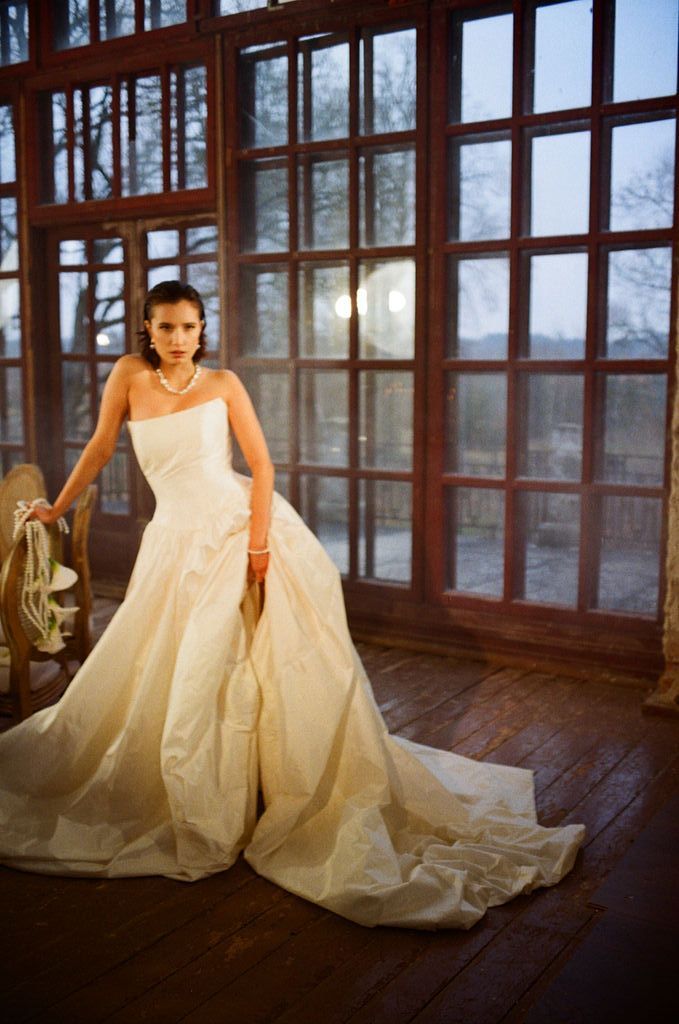 Bride in strapless ivory ballgown with long train poses elegantly before large wooden-framed windows at dusk.