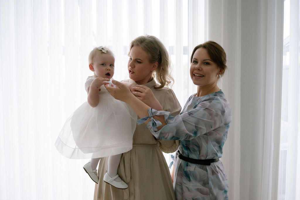 Two women in elegant dresses hold a baby girl in a white dress near a bright window.
